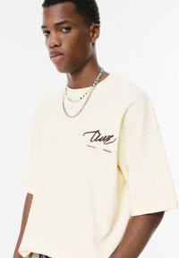 Jasnożółta, oversize t-shirt z krótkimi rękawami, z nadrukowanym czerwonym tekstem i żebrowaną strukturą. Nałożone naszyjniki z koralikami i łańcuchem.