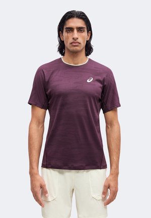 Homme aux cheveux bruns mi-longs portant un t-shirt de sport bordeaux à manches courtes et un short de couleur claire, debout devant un fond clair uni.