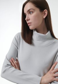 Vrouw met steil bruin haar in een lichtgrijze coltrui, met gekruiste armen, kijkend naar de zijkant tegen een witte achtergrond.