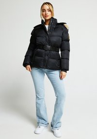 Svart dunjakke med belte, med pelsforet hette, teksturert stoff og knappestengning, kombinert med lyseblå jeans og hvite joggesko.