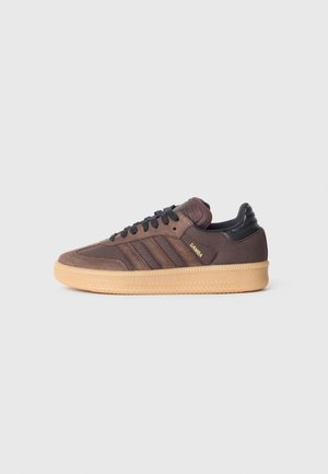 Bruine Adidas Samba sneaker met zwarte veters, zwarte hieltab en een gum rubberen zool op een eenvoudige grijze achtergrond.