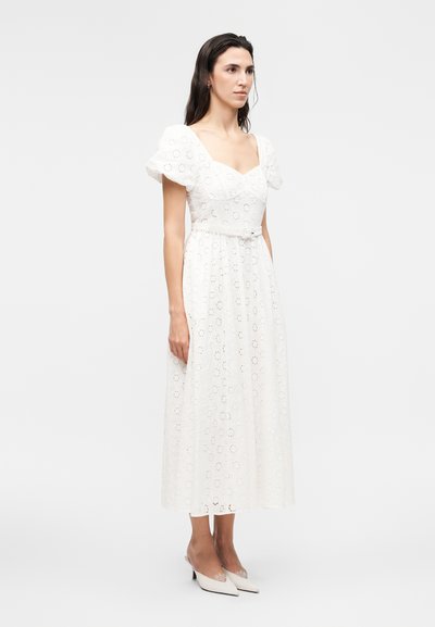 Γυναίκα με μακριά σκουρόχρωμα μαλλιά που φοράει λευκό φόρεμα μέσης με λευκή δαντέλα με τρυπητά μοτίβα, φουσκωτά μανίκια και μυτερά λευκά τακούνια, στέκεται μπροστά σε ένα απλό φόντο.