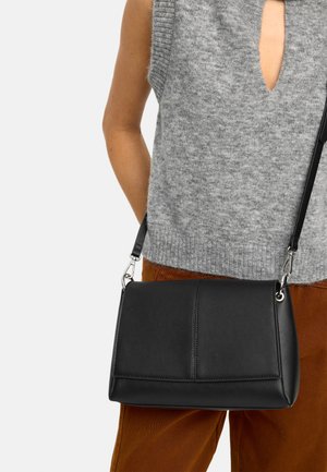 Osoba ubrana w szary sweter bez rękawów i brązowe spodnie, niosąca czarną skórzaną torebkę na ramię z srebrnymi zapięciami.