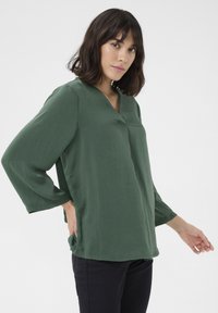 Grøn, langærmet bluse med V-hals. Blødt, let stof med en afslappet pasform. Glat tekstur og sidelommer.