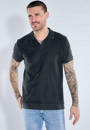 Mann mit kurzen Haaren und Tätowierungen an beiden Armen, trägt ein schwarzes kurzärmeliges Poloshirt und hellblaue Jeans, steht vor hellem Hintergrund.