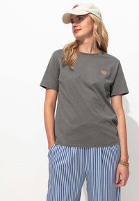 T-shirt en coton gris à manches courtes et col rond, avec un petit logo en forme de cœur doré sur la poitrine. Associé à un pantalon rayé bleu et blanc.