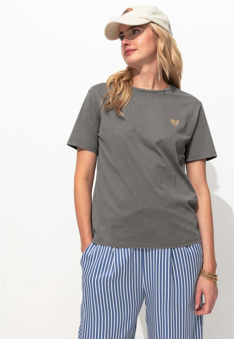 T-shirt en coton gris à manches courtes et col rond, avec un petit logo en forme de cœur doré sur la poitrine. Associé à un pantalon rayé bleu et blanc.