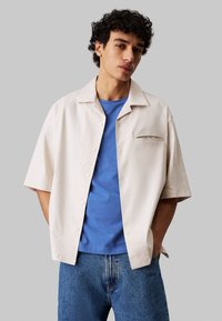 Korte mouw beige button-up shirt met een borstzak, gedragen over een blauwe t-shirt en gecombineerd met blauwe jeans, met een casual pasvorm.