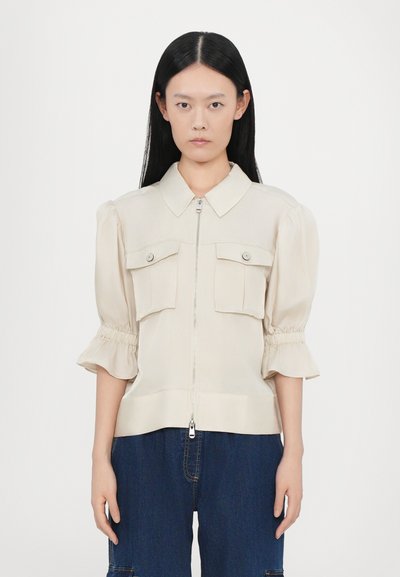 Beige bluse med lynlås foran, korte puffærmer og to brystlommer. Glat stof med subtil glans og sammensyninger ved ærmerne.