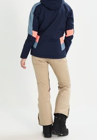 Marineblaue und pastellfarbige, farblich abgesetzte Winterjacke kombiniert mit beigen Skihosen, versehen mit Reißverschlüssen und schwarzen Schneeschuhen. Texturierte Stoffe deutlich erkennbar.