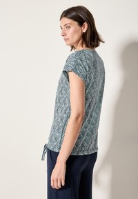 Hladký top s krátkým rukávem v barvě teal se vzorem listů a šňůrkovým detailem v oblasti pasu. Vyrobeno z lehkého a měkkého materiálu.