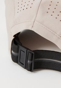 Beige cap with perforated design, nylon strap, and black adjustable buckle. Features a textured finish and contrasting stitching detail.