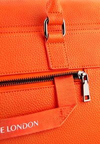 Detalj narančaste teksturirane kožne torbe koja sadrži crni patentni zatvarač i srebrni povlačnik, s brendiranim remenom i kontrastnim šavovima.