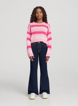 Ragazza con capelli ricci che indossa un maglione a righe rosa e bianco, jeans scuri svasati e scarpe da ginnastica bianche, in piedi davanti a uno sfondo semplice.