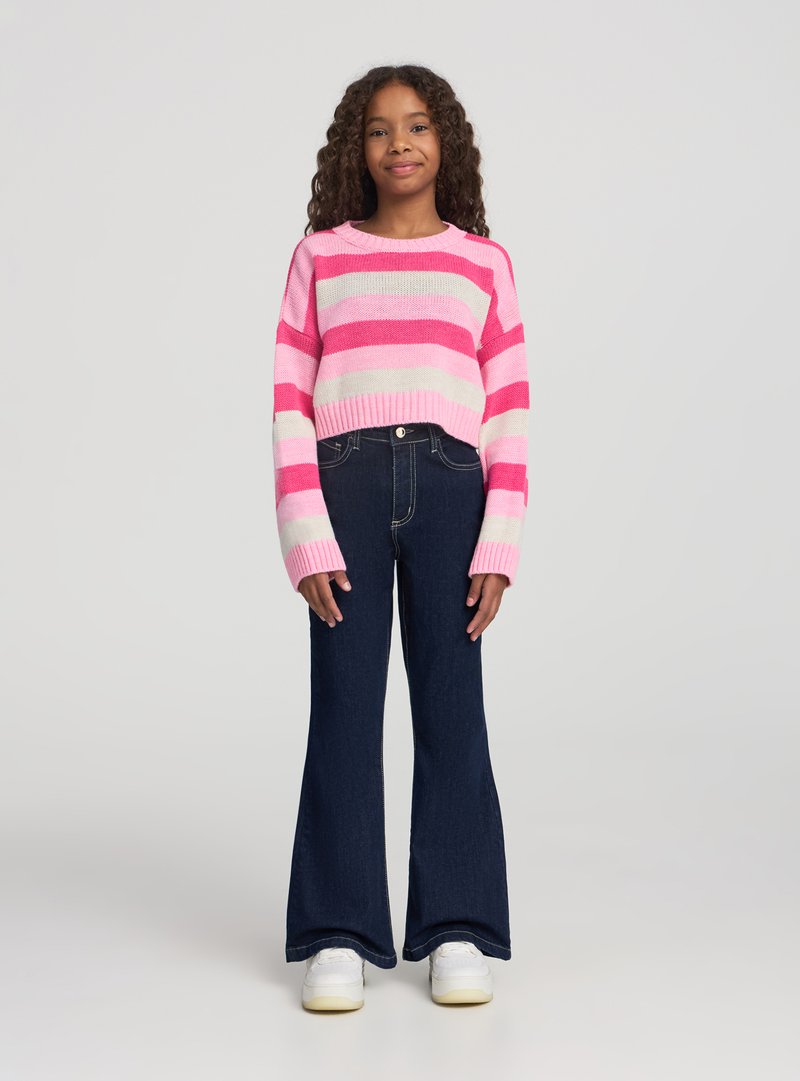 Ragazza con capelli ricci che indossa un maglione a righe rosa e bianco, jeans scuri svasati e scarpe da ginnastica bianche, in piedi davanti a uno sfondo semplice.