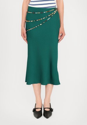 Groene midi-rok van een gladde stof, met een gedrapeerd ontwerp en decoratieve kraal kettingdetail langs de taille. G gedragen met zwarte schoenen.