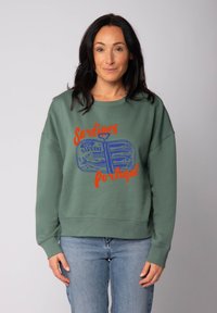 Frau mit langen dunklen Haaren, die einen grünen Pullover mit blauen Sardinen in einer Dose und rotem Text "Sardinen Portugal" trägt, kombiniert mit blauen Jeans.