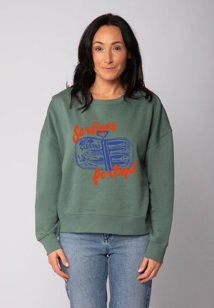 Vrouw met lang donker haar in een groene sweater met blauwe sardines in een blik en rode tekst "Sardines Portugal", gecombineerd met blauwe jeans.