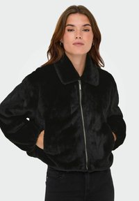 Zwarte nepbontjack met een staande kraag, voorrits en elastische manchetten. Zachte textuur en losse pasvorm, gecombineerd met zwarte broek.