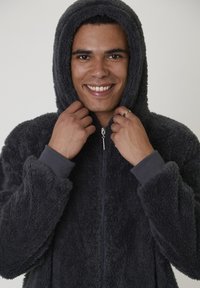 Homme souriant en mettant la capuche d'une veste à fermeture éclair en peluche gris foncé sur sa tête, portant une bague en or à sa main gauche.