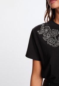 Zwarte katoenen t-shirt met een decoratief patroon van zilveren rhinestones, die in bloemige ontwerpen zijn gerangschikt over de linkerschouder en het borstgebied.