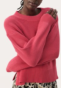Różowy sweter dzianinowy o luźnym kroju, z prążkowanymi mankietami i dołem. Posiada teksturowane wzory oraz boczne rozcięcia. Nosi się go na spodniach w panterkę.