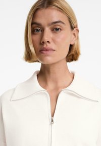 White knit sweatshirt featuring a collared, zippered neckline and ribbed texture. Simple design and soft fabric with a clean finish.