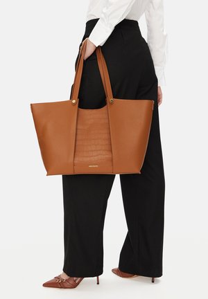 Mujer con camisa blanca y pantalones negros sosteniendo un gran bolso tote de cuero marrón con panel central texturizado y asas dobles.