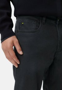 Donkergrijze jeans met een gestructureerde afwerking, voorzien van een voorzak met een kleine gele accent en een zichtbare knopenfly. Hand rustend aan de zijkant.