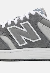 Sapatilhas de camurça e couro cinzas com atacadores brancos, apresentando um proeminente logo "N" cinza na lateral e detalhes texturizados na entressola.