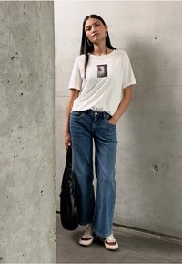 Camiseta blanca con un gráfico en la parte frontal, combinada con jeans azules holgados. Una bolsa de mano negra y zapatillas ligeras con acentos rojos completan el look.