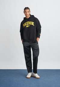Čierna mikina s kapucňou a žltým logom "Reebok" na prednej strane, doplnená čiernymi teplákovými nohavicami. Biele tenisky s hnedými podrážkami. Jednoduché pozadie.