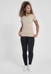 Beżowy, dopasowany, krótki rękaw z prążkowaną fakturą, zestawiony z czarnymi, prążkowanymi legginsami. Modelka ma na sobie białe trampki, stoi na neutralnym tle.