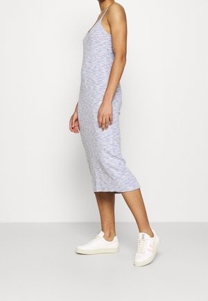 Femme portant une robe midi sans manches bleu clair et des baskets blanches, debout de profil devant un fond clair uni.