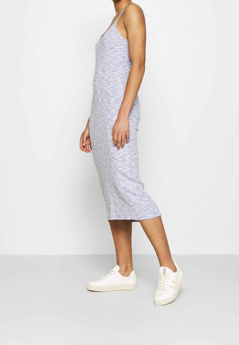 Femme portant une robe midi sans manches bleu clair et des baskets blanches, debout de profil devant un fond clair uni.