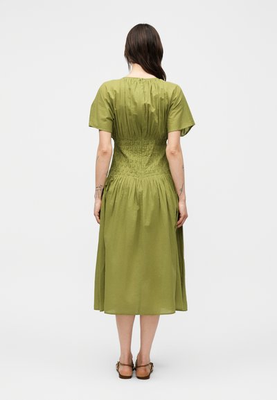 Γυναίκα με μακριά σκούρα μαλλιά που φοράει ένα φορεματάκι μήκους μέχρι τα γόνατα σε ελιά πράσινο χρώμα με κοντά μανίκια και σφηκοειδή μέση, στέκεται με την πλάτη γυρισμένη, σε λευκό φόντο.