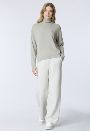 Maglione a collo alto grigio chiaro con una texture morbida, abbinato a pantaloni ampi color crema e sneakers bianche. Logo notevole sul maglione.