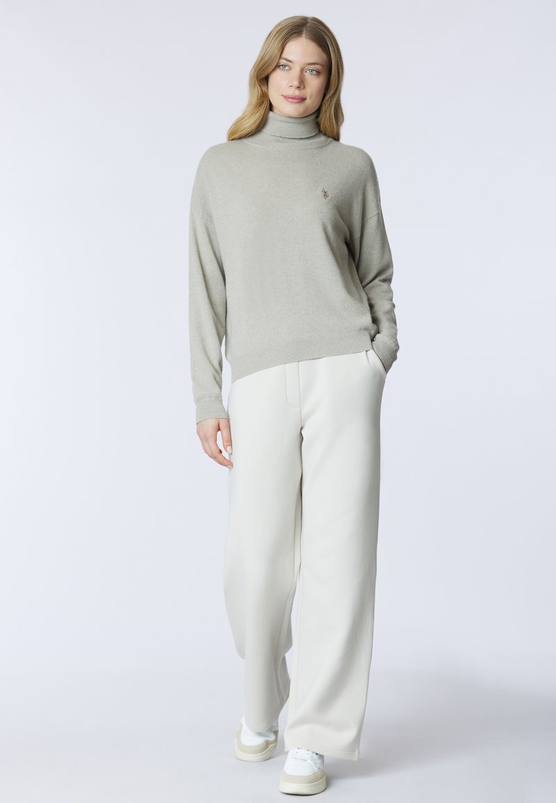 Maglione a collo alto grigio chiaro con una texture morbida, abbinato a pantaloni ampi color crema e sneakers bianche. Logo notevole sul maglione.