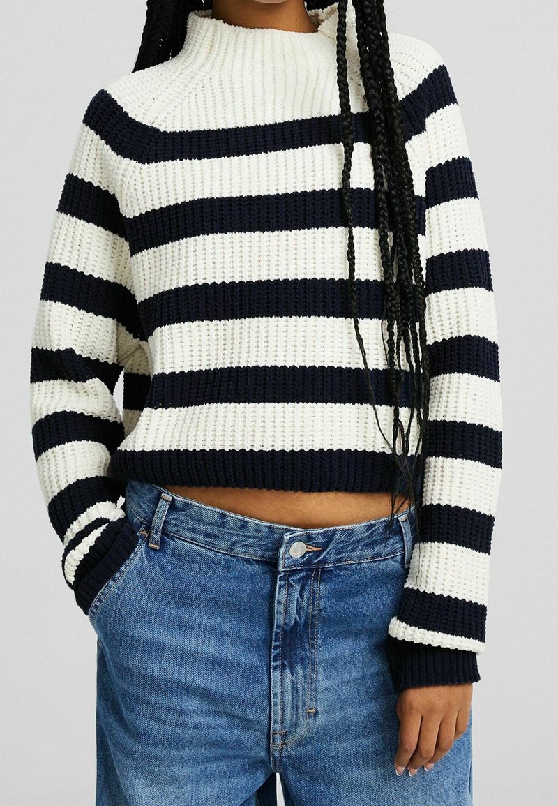Pletený svetr s navy a krémovými vodorovnými pruhy, vysokým límcem a žebrovanými manžetami. Spárován s vysoce pasovými bledě modrými džíny.