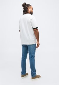 Άνδρας με ντραντλoκς, φοράει λευκό μπλουζάκι με μαύρη περιγραφή, μπλε τζιν και καφέ αθλητικά παπούτσια, στέκεται έχοντας την πλάτη γυρισμένη σε απλό λευκό φόντο.