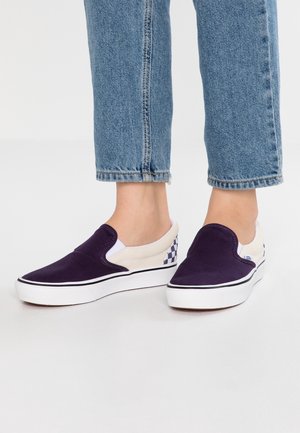 Stopy w fioletowo-białych trampkach typu slip-on z czarno-białym wzorem w kratkę na piętach i niebieskich dżinsach.
