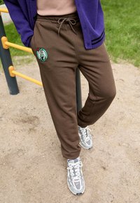 Pantaloni della tuta marroni in morbido tessuto, con logo ricamato. Vita con cordino regolabile e vestibilità comoda. Abbinati a sneakers grigie.