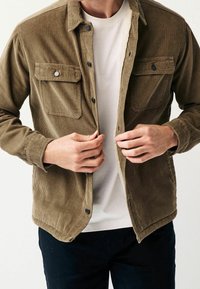 Braune Cordjacke aus Cord mit strukturierter Oberfläche, Druckknöpfen, zwei Brusttaschen und einem Futterdetail. Trägt man über einem weißen T-Shirt.