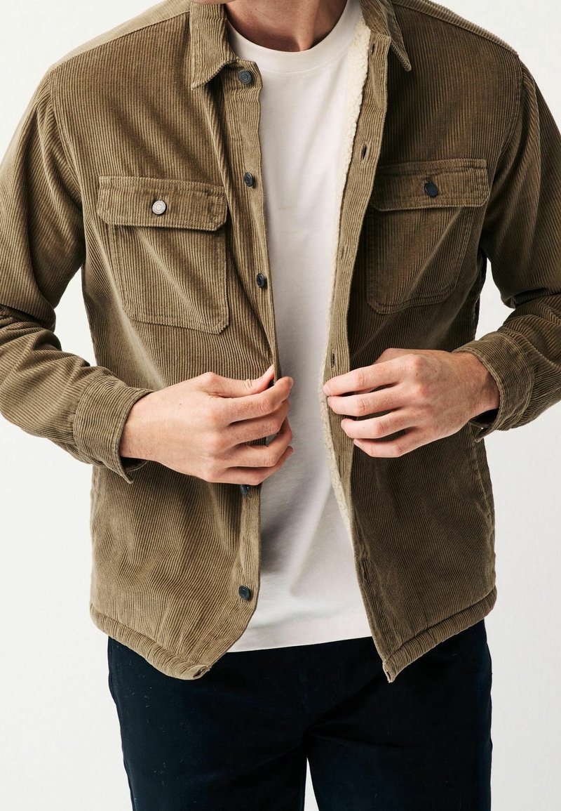 Braune Cordjacke aus Cord mit strukturierter Oberfläche, Druckknöpfen, zwei Brusttaschen und einem Futterdetail. Trägt man über einem weißen T-Shirt.