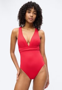 Γυναίκα με σγουρά μαλλιά που φοράει ένα αμάνικο κόκκινο ολόσωμο μαγιό με βαθύ V στο ντεκολτέ και ζώνη με ραβδώσεις, στεκόμενη μπροστά σε λευκό φόντο.