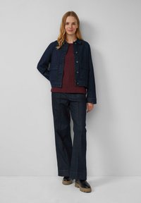 Vrouw die tegen een lichtgrijze muur staat, gekleed in een donkerblauwe jas, een kastanjebruine trui, donkere wijde broek en zwarte laarzen met beige zolen.
