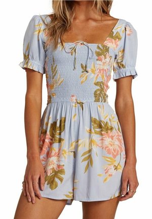 Femme portant une combinaison à fleurs bleu clair avec des manches courtes bouffantes, un corsage smocké, un décolleté carré et un détail à nouer au centre de la poitrine.