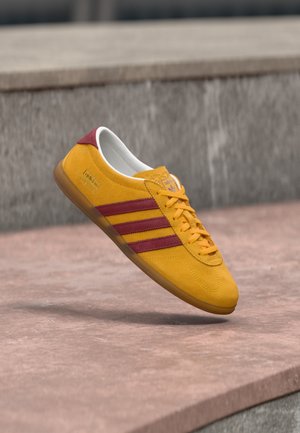 Gele suède sneaker met rode strepen, platte zool en gevoerde kraag; beschikt over een vetersluiting en een merklabel.