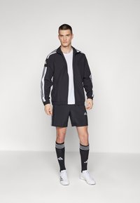 Zwarte sportjack met witte strepen, bijpassende zwarte shorts en kniehoge zwarte sokken met witte accenten. Witte sportschoenen.