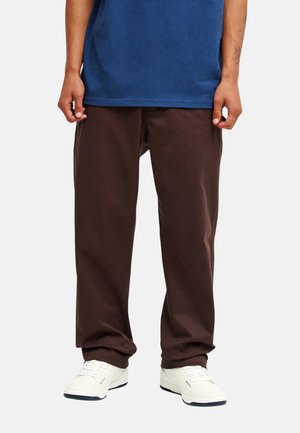 Person trägt lockere dunkelbraune Hose, weiße Sneakers und ein blaues Hemd, steht vor einem einfarbigen hellen Hintergrund.